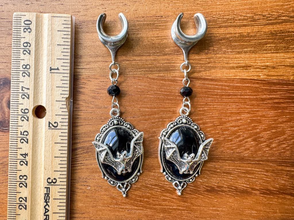 Silver and black bat crest plug earrings with gothic bat dangles for stretched ears next to ruler for sizing.